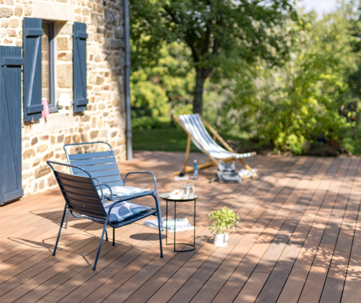 Réalisation d'une terrasse composite Atmosphère Nuances IPE de Silvadec, au bord d'une maison en pierre avec des volets bleu marine. Deux chaises de jardin sont présente ainsi qu'une petite table avec deux verres dessus. 