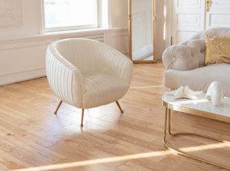 Salon illuminé, avec un parquet massif clair. Un petit fauteuil, un canapé et une table blanche sont présents avec les pieds de couleur or.